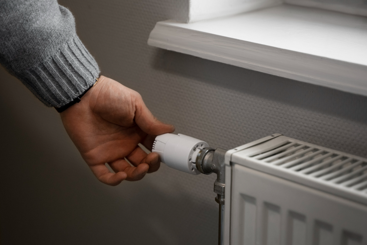 Main ajustant le thermostat d’un radiateur blanc situé sous une fenêtre.